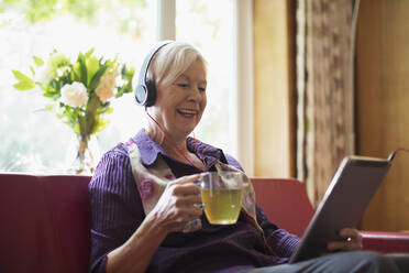 Happy senior woman with headphones and digital tablet drinking tea - CAIF29862