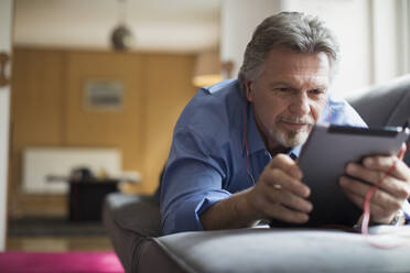 Senior man using headphones and digital tablet on living room sofa - CAIF29875