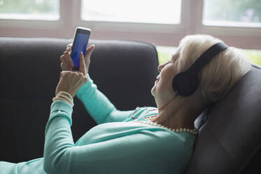 Senior woman relaxing with headphones and smart phone on sofa - CAIF29876