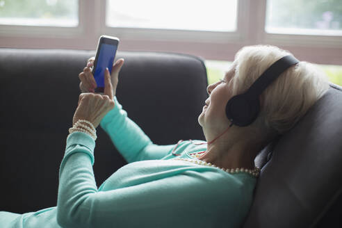 Ältere Frau entspannt sich mit Kopfhörern und Smartphone auf dem Sofa - CAIF29876