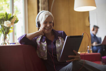 Glückliche ältere Frau benutzt Kopfhörer und digitales Tablett auf dem Sofa - CAIF29877