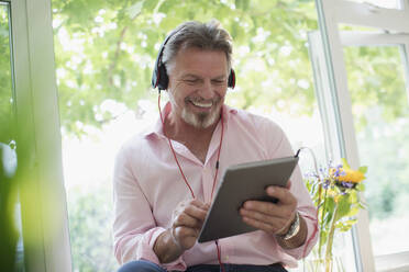 Glücklicher älterer Mann mit Kopfhörer und digitalem Tablet am Fenster - CAIF29879