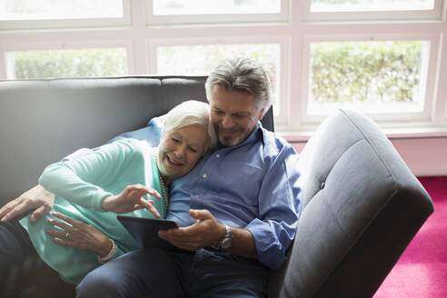 Glückliches Seniorenpaar kuschelt und benutzt digitales Tablet auf dem Sofa - CAIF29880
