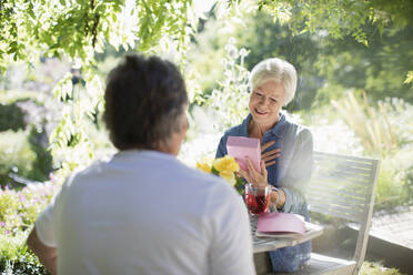 Glückliche ältere Frau öffnet Geschenk von Ehemann auf sonniger Sommerterrasse - CAIF29883