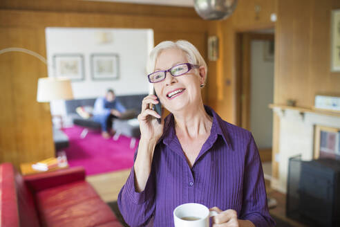 Glückliche ältere Frau mit Kaffee, die sich mit einem Smartphone im Wohnzimmer unterhält - CAIF29915