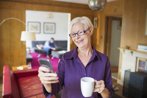 Glückliche ältere Frau benutzt Smartphone und trinkt Tee im Wohnzimmer - CAIF29919