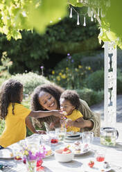 Glückliche Mutter und Töchter essen an einem sonnigen Sommer-Gartentisch - CAIF29987