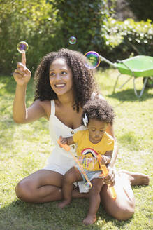 Happy mother and toddler daughter blowing bubbles in sunny grass - CAIF29993