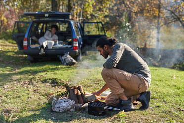Mann bereitet Lagerfeuer vor dem Auto im sonnigen Herbstfeld - CAIF30027