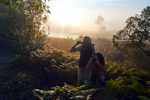 Neugieriges junges Paar wandert mit Fernglas und Kamera in sonnigen Wäldern - CAIF30050