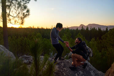 Junger Mann mit Verlobungsring macht der Frau bei Sonnenuntergang einen Heiratsantrag auf einem Felsen - CAIF30054