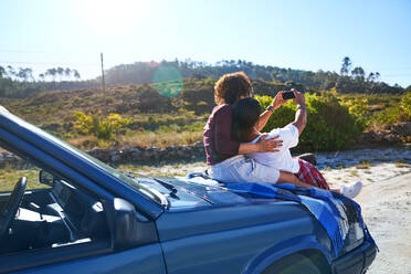 Carefree young couple taking selfie on hood of car at sunny roadside - CAIF30060