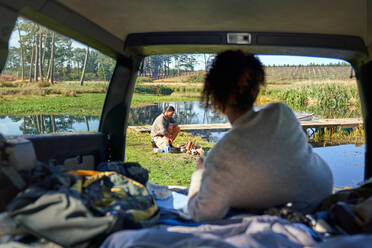 Young woman in car watching boyfriend prepare campfire - CAIF30065