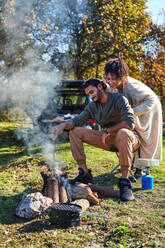 Happy young couple enjoying campfire in sunny autumn field - CAIF30068