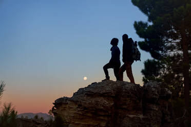 Silhouette young hiker couple enjoying view of moon from rock at dusk - CAIF30069