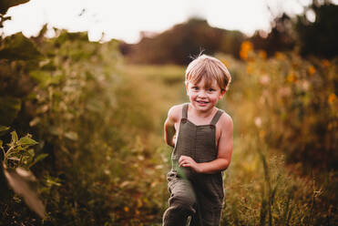 Happy boy running in a flower field wearing green dungarees - CAVF90174