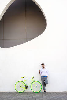 Young bearded man leaning against a white wall using the mobile phone - CAVF90199