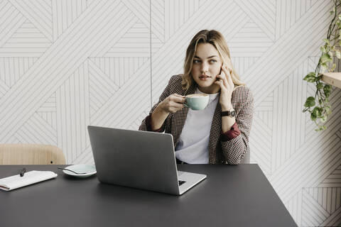 Junge Frau, die mit ihrem Smartphone telefoniert und Kaffee trinkt, während sie in einem Café sitzt, lizenzfreies Stockfoto