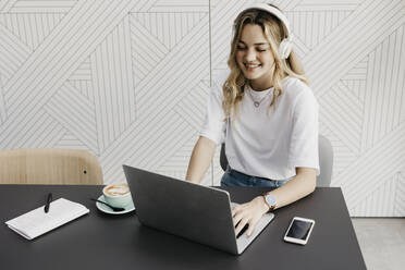 Smiling woman wearing headphones working on laptop sitting in cafe - LHPF01300
