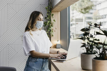 Frau mit Gesichtsschutzmaske, die einen Laptop benutzt, während sie in einem Café steht (Covid-19) - LHPF01307