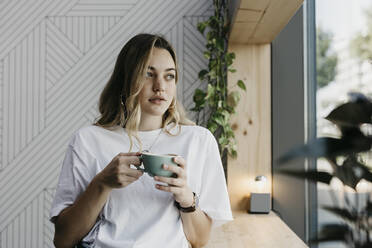Junge Frau hält eine Kaffeetasse und schaut durch ein Fenster, während sie in einem Café steht - LHPF01309