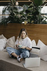 Young woman holding coffee cup while using laptop sitting on sofa - LHPF01314