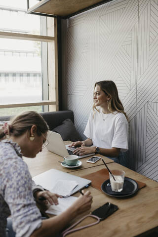 Young woman using laptop while female friend writing sitting in cafe stock photo