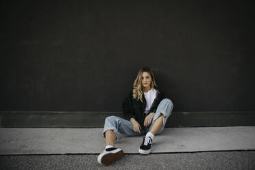 Young woman sitting on footpath against wall - LHPF01327