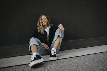 Young woman smiling while sitting on footpath against wall - LHPF01328