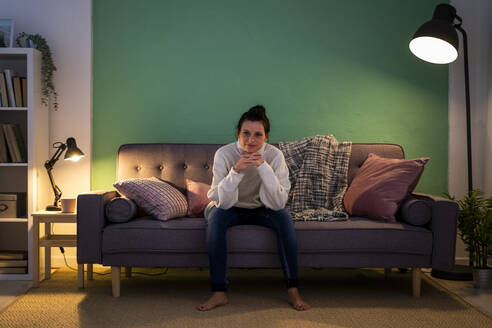 Lächelnde Frau mit Hand am Kinn, die zu Hause auf der Couch sitzt - GIOF09294