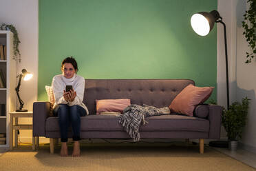 Frau benutzt Smartphone und schaut weg, während sie zu Hause auf dem Sofa sitzt - GIOF09298