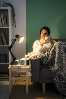 Mittlere erwachsene Frau hält Kaffeetasse, während sie zu Hause auf dem Sofa sitzt - GIOF09306
