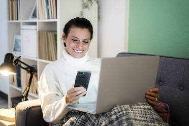 Lächelnde Frau benutzt Smartphone und Laptop, während sie zu Hause sitzt - GIOF09308