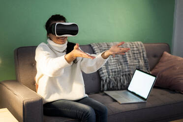 Mittlere erwachsene Frau trägt einen virtuellen Simulator, während sie zu Hause auf dem Sofa sitzt - GIOF09312