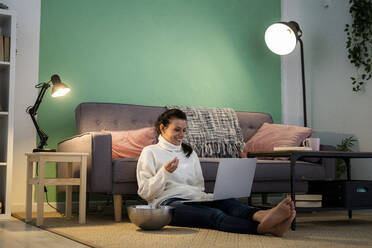 Mittlere erwachsene Frau, die einen Laptop benutzt, während sie zu Hause auf dem Boden sitzt und isst - GIOF09316