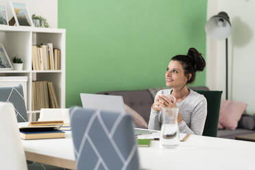 Mid adult woman looking away while holding coffee cup sitting on chair at home - GIOF09318