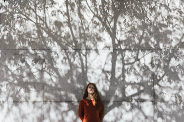 Young woman with hands behind back standing against tree shadow wall - TCEF01211