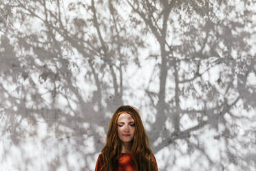 Redhead woman standing with eyes closed against tree shadow wall - TCEF01212