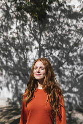Young redhead woman standing with eyes closed against tree shadow wall - TCEF01213
