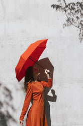 Junge Frau in rotem Kleid, die einen Regenschirm hält, während sie vor einer Schattenwand steht - TCEF01218