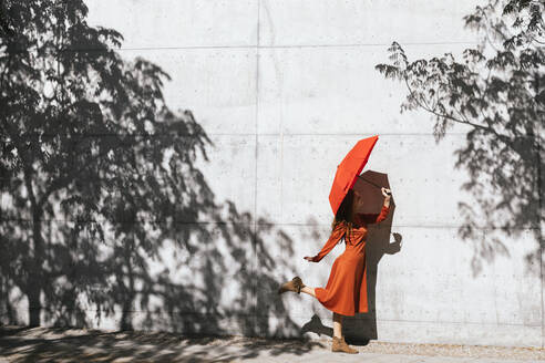 Frau in rotem Kleid, die einen roten Regenschirm hält, während sie vor einer Baumschattenwand steht - TCEF01219
