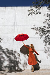 Junge Frau in rotem Kleid hält fliegenden Regenschirm, während sie gegen einen Baum Schattenwand steht - TCEF01221