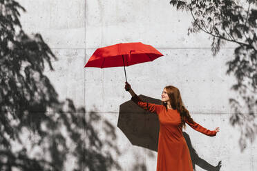 Lächelnde rothaarige Frau, die einen Regenschirm hält, während sie vor einer Baumschattenwand steht - TCEF01222