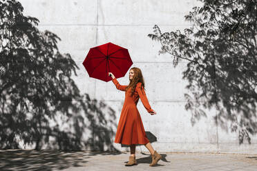 Frau in rotem Kleid, die einen roten Regenschirm hält, während sie vor einer Baumschattenwand steht - TCEF01223