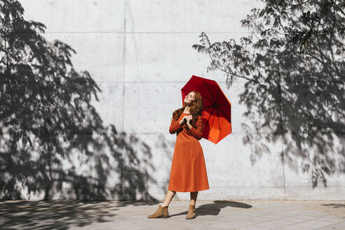 Lächelnde Frau, die einen Regenschirm hält, während sie vor einer Baumschattenwand steht - TCEF01224