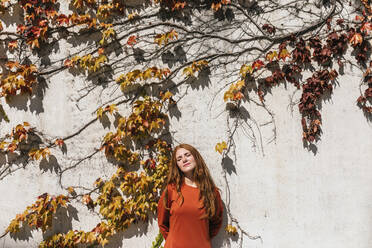 Woman with eyes closed and hands behind back standing against ivy wall  - TCEF01230