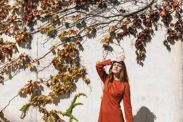 Woman with hand on head standing against ivy wall - TCEF01233