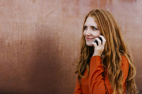 Smiling woman talking on mobile phone by brown wall - TCEF01258