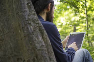 Älterer Mann benutzt digitales Tablet und lehnt sich an einen Baum - MAMF01382