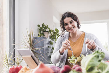 Smiling woman eating salad while using digital tablet home - UUF21938